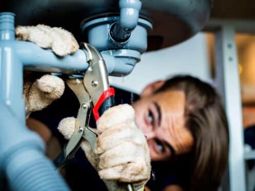 A plumber wearing gloves tightens a pipe connection under a kitchen sink using pliers, ensuring a secure plumbing repair in a Dallas home.