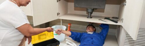 Plumber in blue uniform fixing kitchen sink pipes while homeowner assists in Dallas home