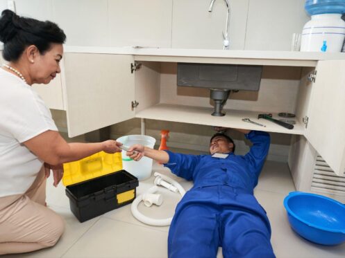 Plumber in blue uniform fixing kitchen sink pipes while homeowner assists in Dallas home